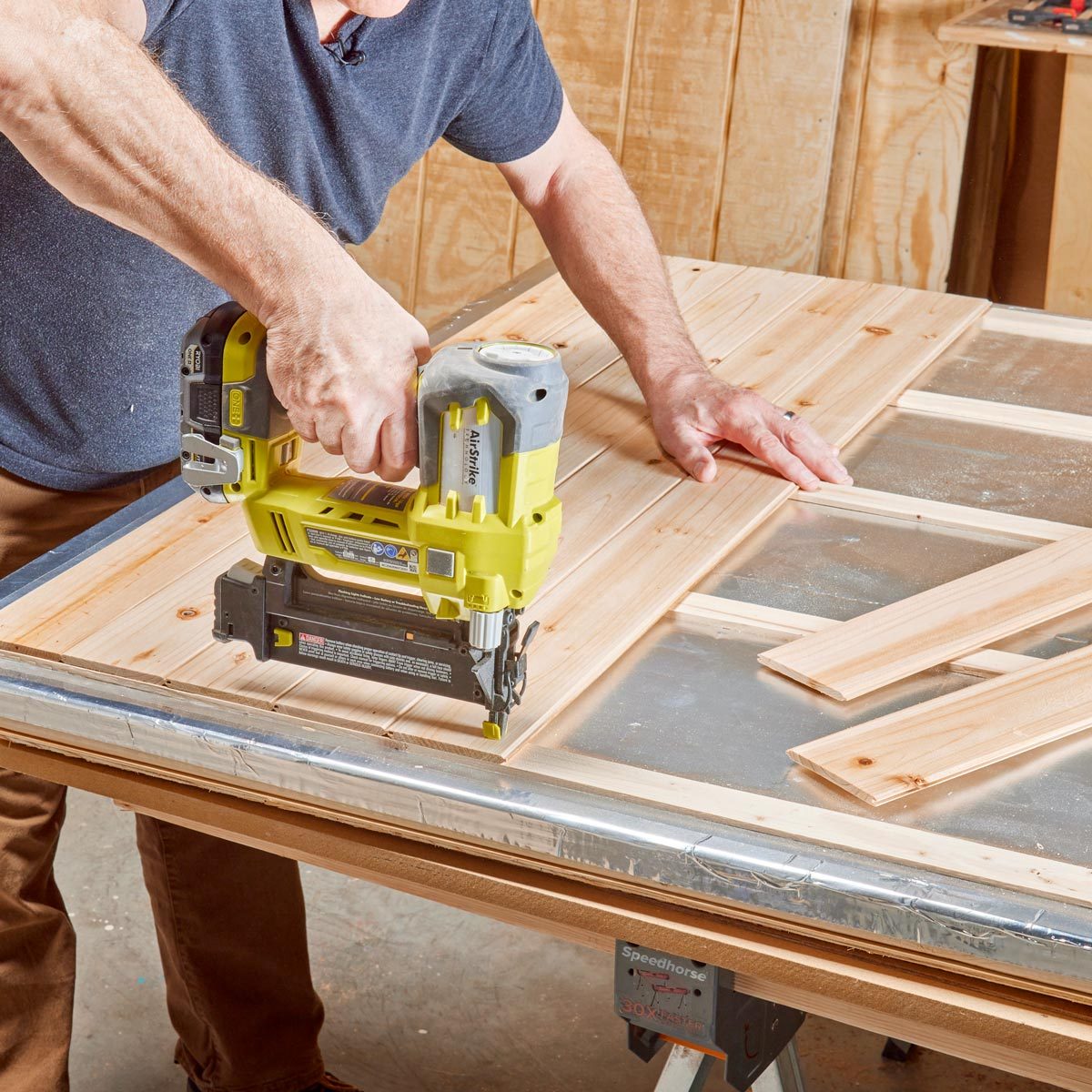 A person uses a nail gun on wooden panels placed on a workbench in a workshop, surrounded by a wooden backdrop and tools.