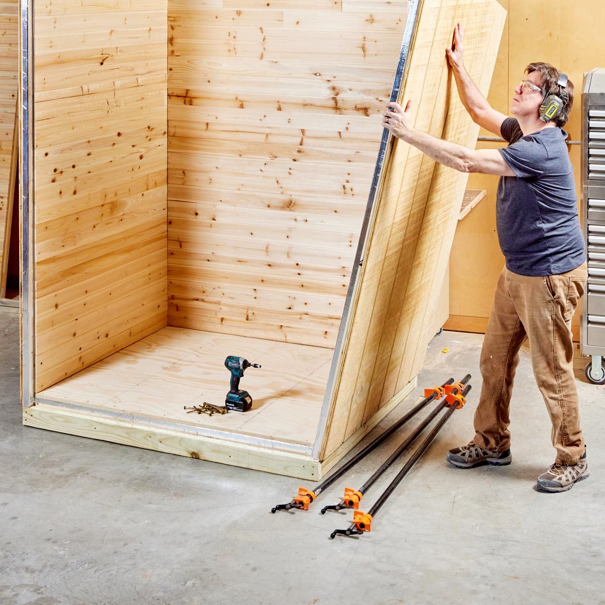 A person adjusts a wooden panel while building a structure in a workshop, surrounded by tools, clamps, and an unfinished wooden frame.