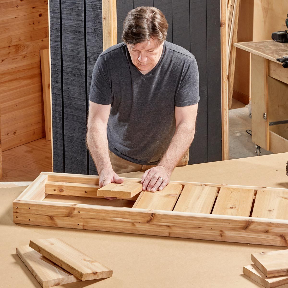 A man places wood pieces into a partially assembled wooden frame on a workbench surrounded by tools and wooden planks in a workshop.