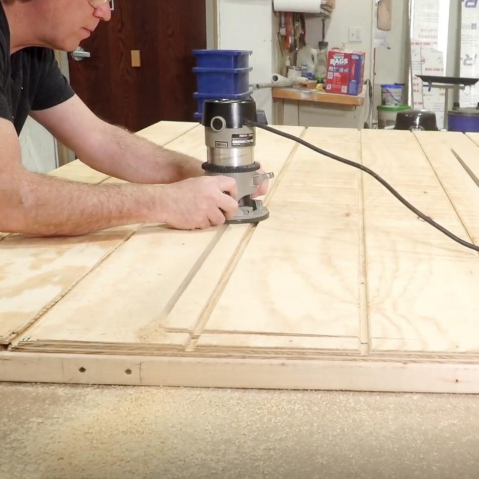 A person uses a router to carve grooves into a large wooden panel on a workbench, surrounded by tools and a workshop environment.