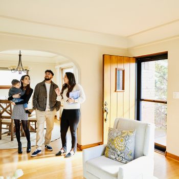 A family tours a bright living room, discussing with a guide while admiring the space. Wooden furniture and a large window add warmth to the environment.