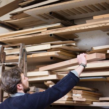 A person is reaching for a wooden plank on a high shelf in a workshop filled with stacked lumber and tools.