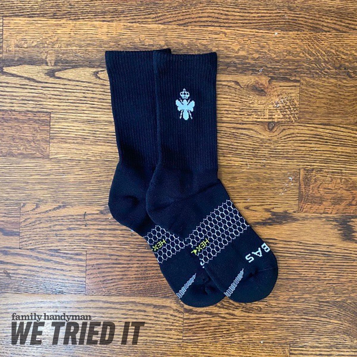 Black socks are displayed horizontally, featuring a logo and textured pattern at the bottom, resting on a wooden surface.