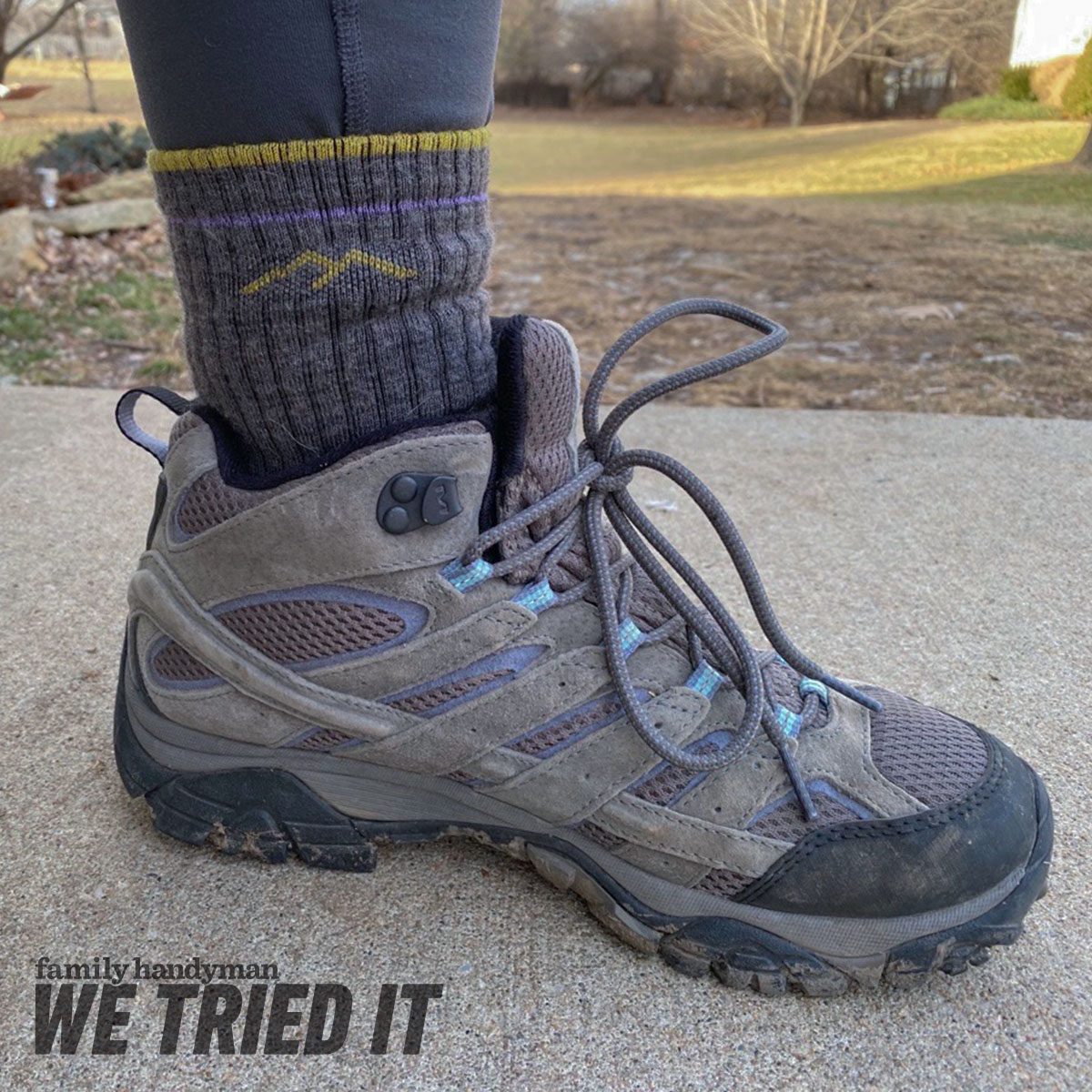 A brown hiking boot laced loosely rests on a concrete surface. The boot is paired with a dark sock featuring a mountain design, with a grassy area in the background.