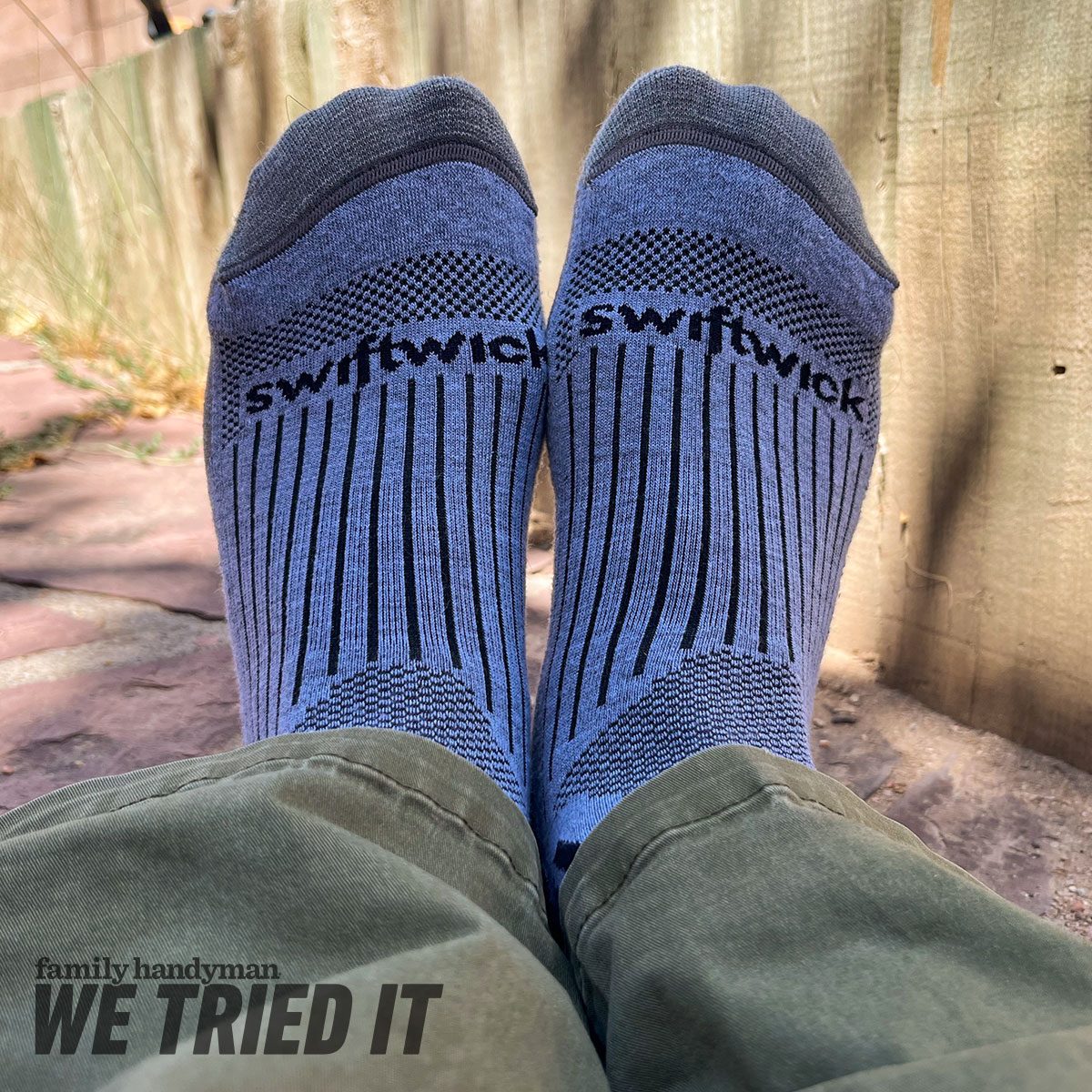 Blue Swiftwick socks are worn by a person seated outdoors on a stone surface, with a wooden fence and grass in the background.