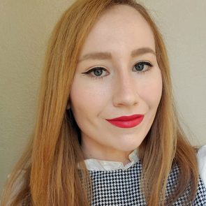A person with long, straight, light brown hair smiles, showcasing red lipstick and a collared outfit with a houndstooth pattern, against a neutral background.