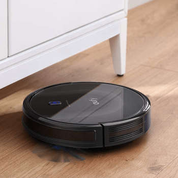 A robotic vacuum cleaner moves across wooden flooring, cleaning beneath a white cabinet, showcasing a sleek black design with a blue indicator light.
