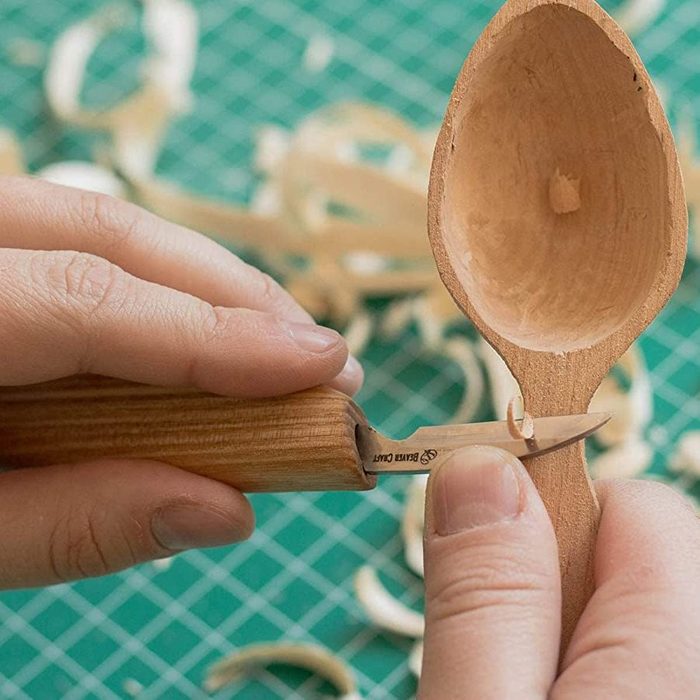 A person carves a wooden spoon using a carving knife, surrounded by wood shavings on a green cutting mat with a grid pattern.