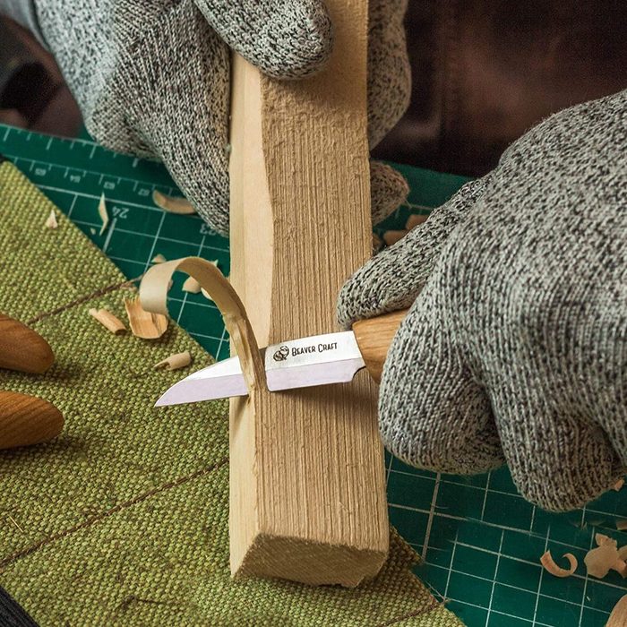 A person uses a carving knife to shape a piece of wood, surrounded by wood shavings on a green mat and a cutting board.