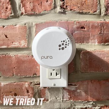 A white device is mounted on a brick wall, plugged into an outlet, suggesting it dispenses fragrance in the surrounding environment.