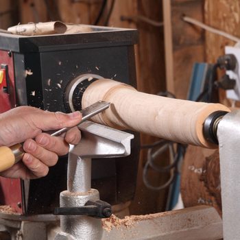 A hand uses a chisel to shape a spinning piece of wood on a lathe, while wood shavings fly in a workshop setting.