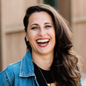 A woman with long, curly hair smiles broadly, showcasing her teeth, while wearing a denim jacket. The background features soft brown tones.