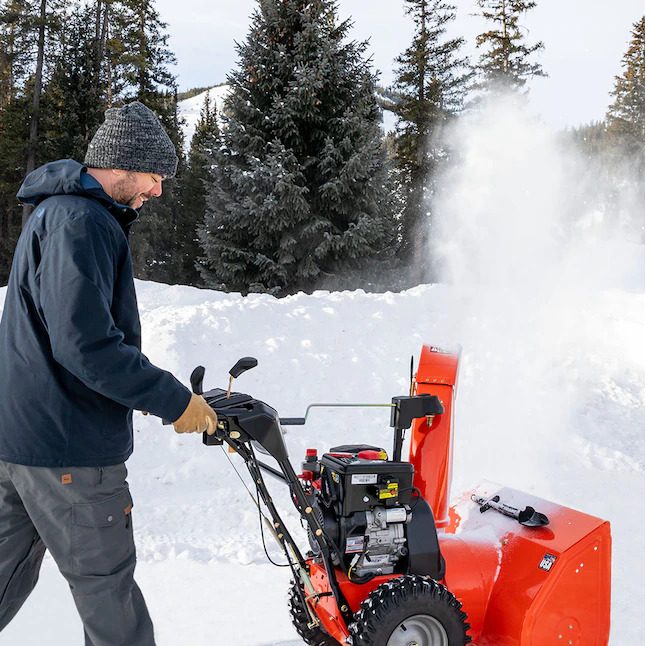 A man operates a snowblower, clearing snow in a winter landscape surrounded by evergreen trees under a clear sky.