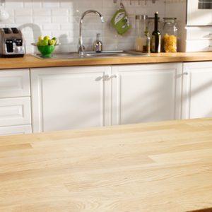 A wooden kitchen countertop holds a bowl of green apples. Behind it, a sink, toaster, and jars are arranged on white cabinetry against a bright wall.