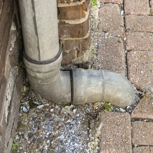 A gray pipe bends at a corner, draining water near a brick wall onto a paved surface, scattered with debris and small plants.