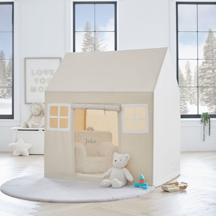 A beige playhouse stands indoors, inviting play. A plush teddy bear sits outside, and a small chair inside is labeled 