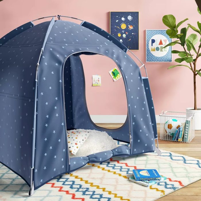 A blue star-patterned tent stands in a brightly colored room, with a soft interior and playful decorations on the walls, surrounded by toys and a plant.