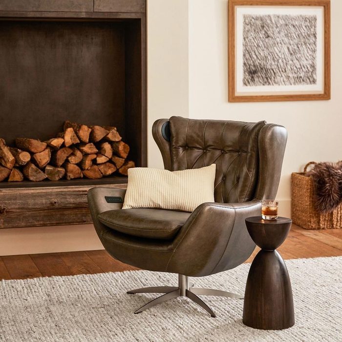 A stylish brown armchair sits beside a wooden side table with a drink, facing a cozy fireplace stacked with logs, on a light rug.