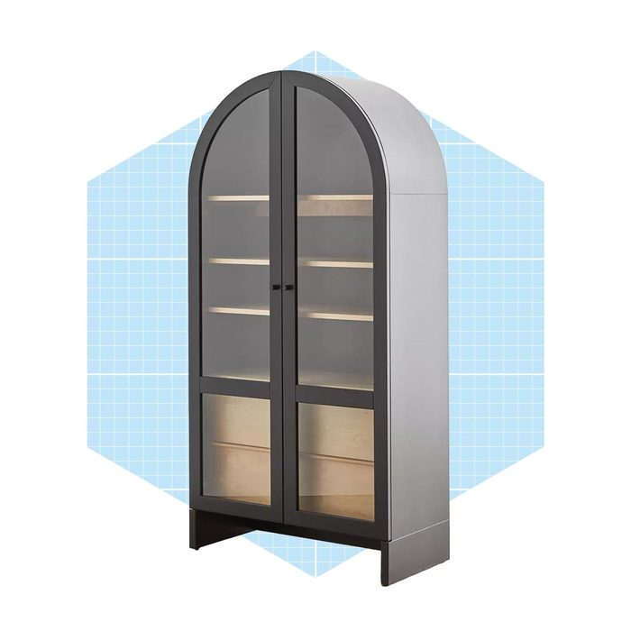 A tall, arched cabinet displays open shelves behind glass doors, featuring a sleek gray finish against a light blue grid background.
