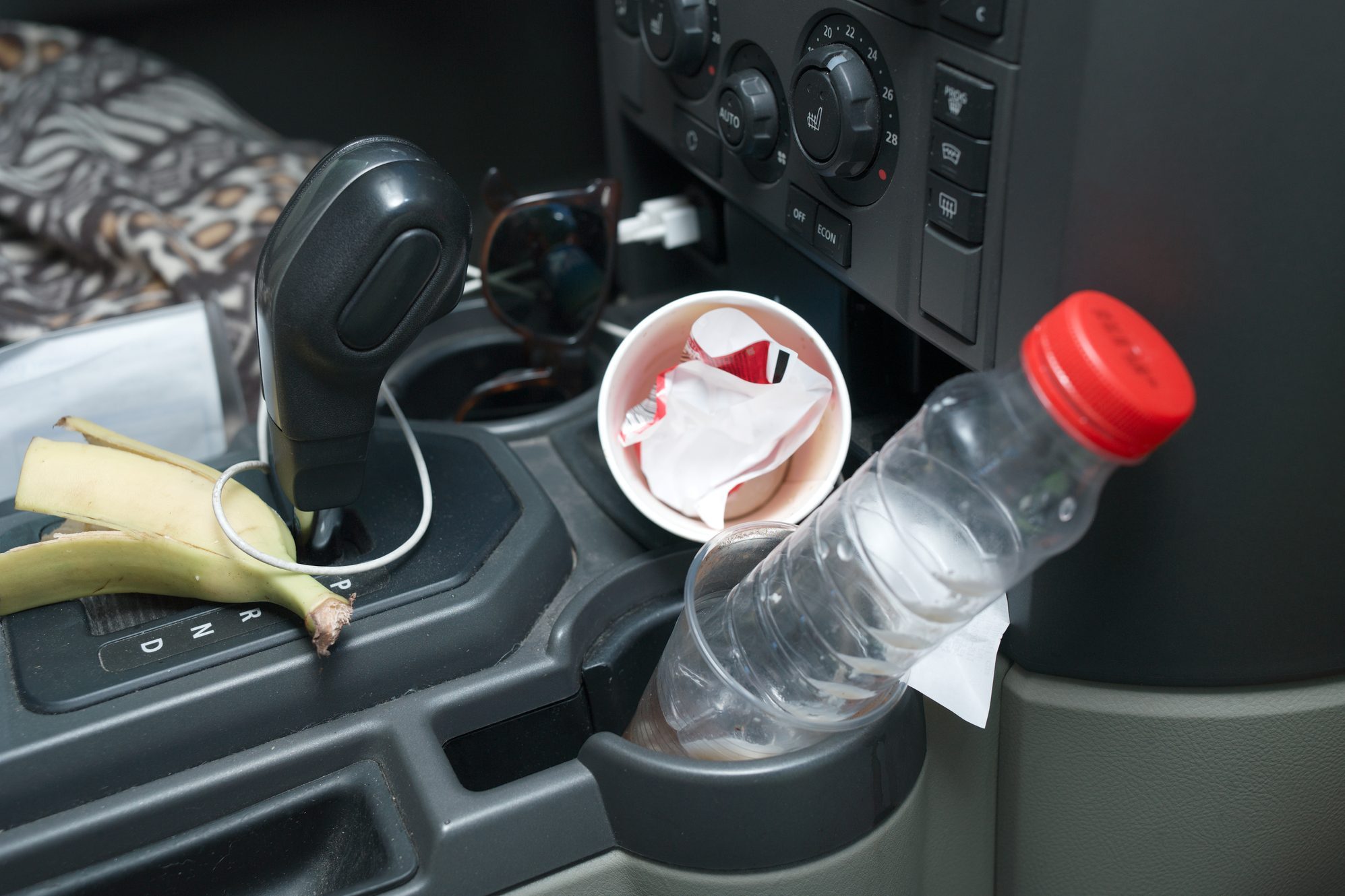 A car's console features a gear shift, a banana, a crumpled cup, and a water bottle, amidst a patterned cloth and sunglasses nearby.