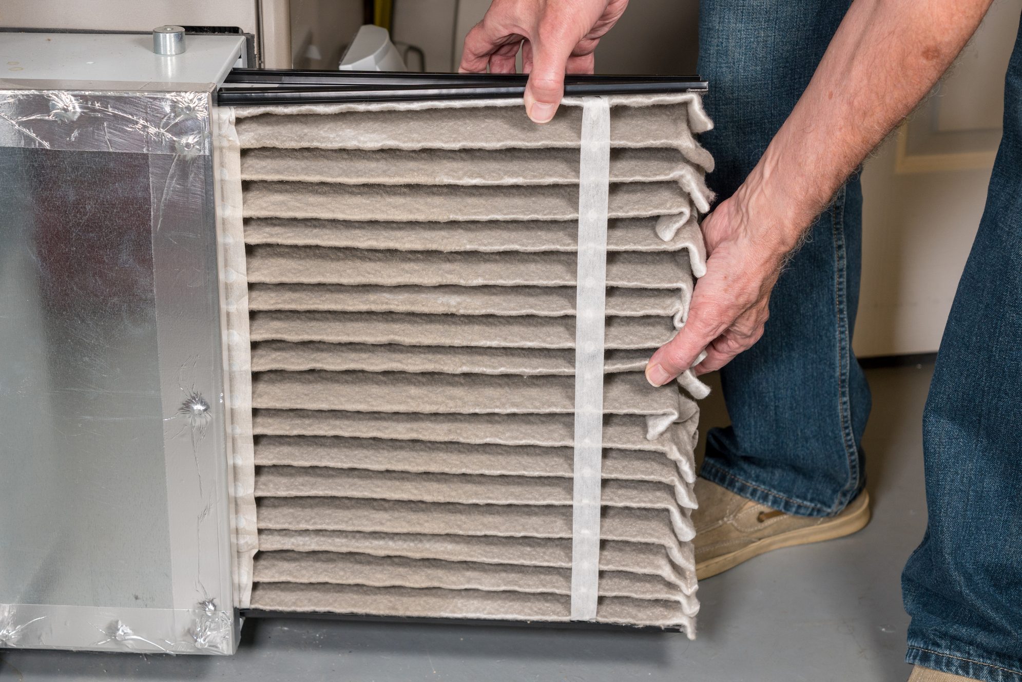 A person removes a thick, folded air filter from a metal housing in a home environment, preparing for maintenance or replacement.