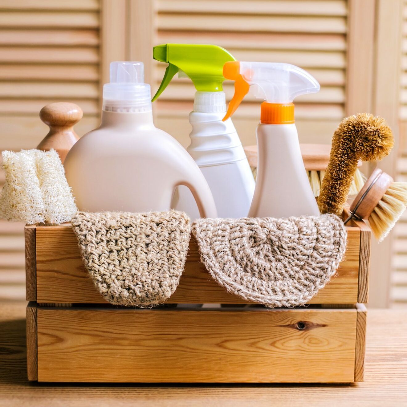 A wooden crate holds various cleaning supplies, including bottles and brushes, surrounded by towels, set against a backdrop of light-colored wood paneling.