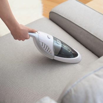 A handheld vacuum cleaner is being used to clean a gray sofa cushion in a bright, modern living room with wooden floors and a small potted plant nearby.