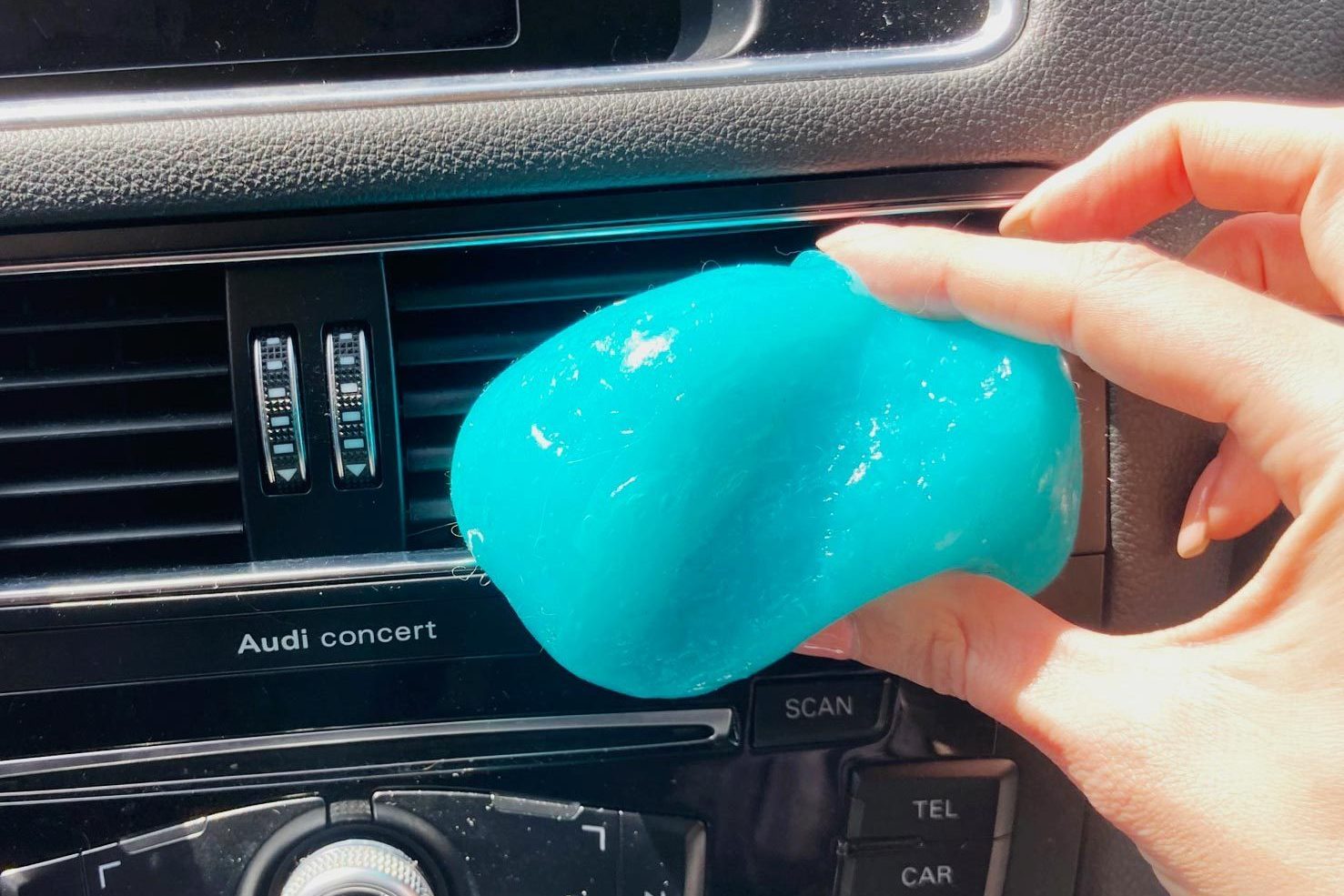A hand holds bright blue slime, pressing it against car air vents, with an Audi concert display and control knobs visible in the dashboard.