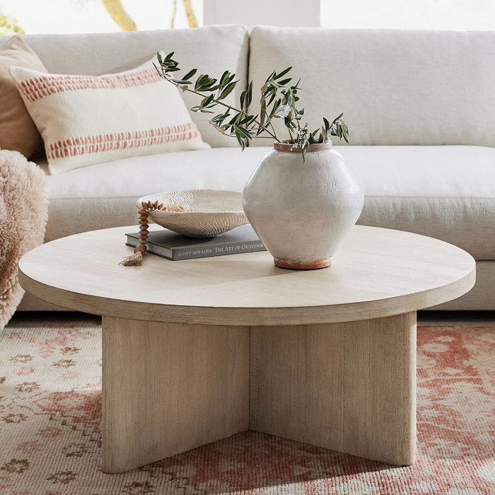 A round wooden coffee table displays a vase with greenery, a bowl, and stacked books in a cozy living room with a light-colored sofa.