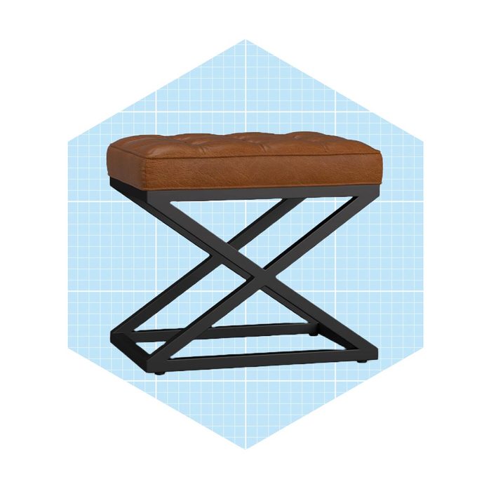 A brown leather stool with a tufted top rests on a black, crisscross metal frame, set against a light blue grid background.