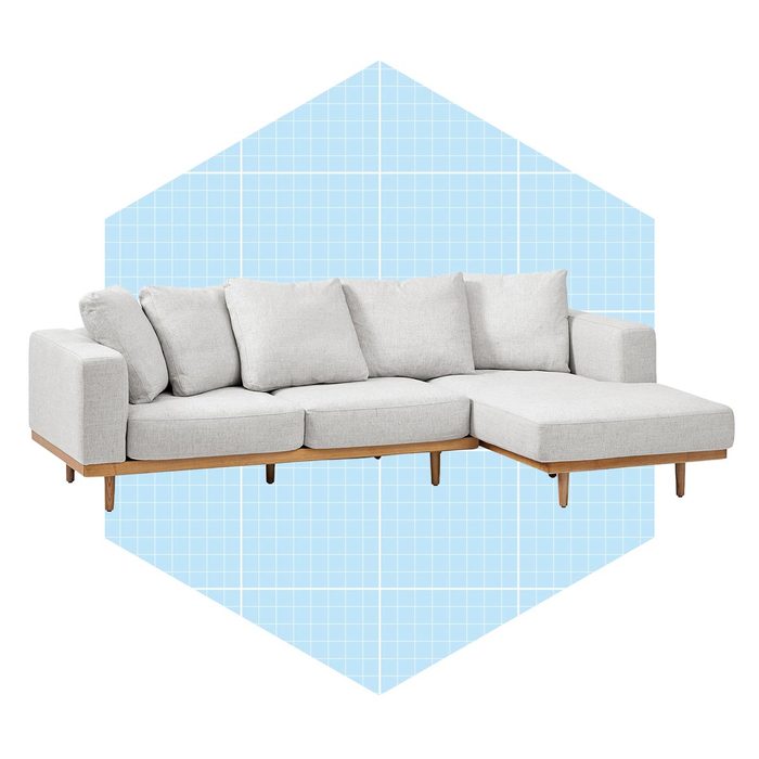 A light gray sectional sofa with plush cushions rests on wooden legs, set against a blue grid-patterned background resembling a house shape.