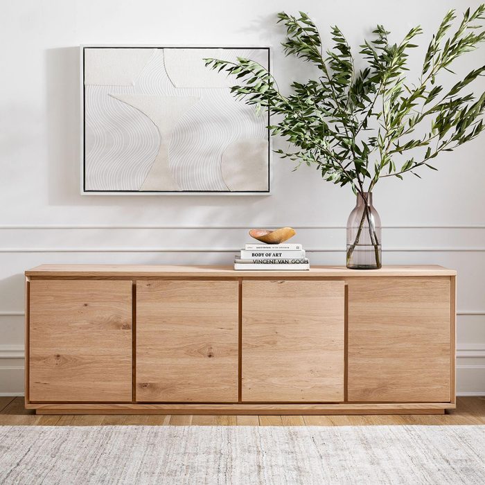 A wooden console table displays stacked books and a fruit bowl, while a vase with greenery stands beside an abstract wall art piece in a bright room.