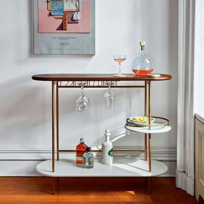 A modern bar cart displays drinks and glassware alongside a framed artwork against a light wall, illuminated by natural light from a nearby window.