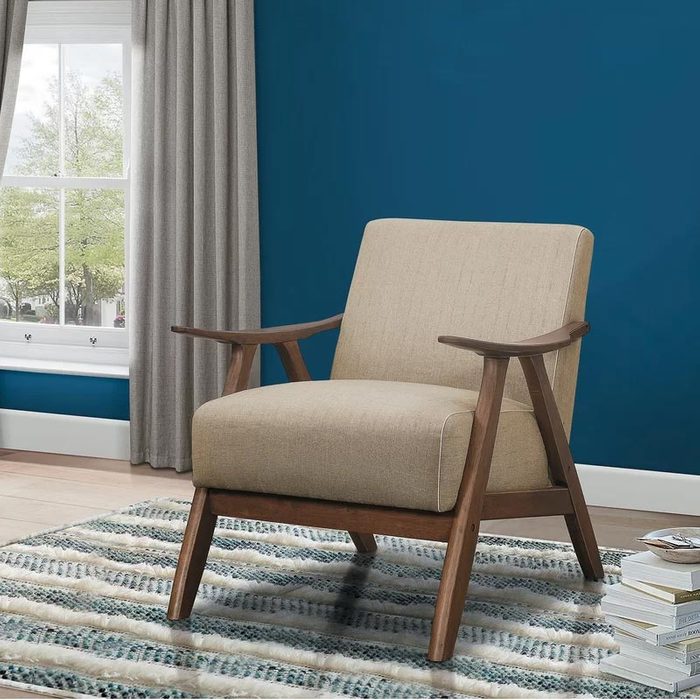 A beige armchair rests on a patterned rug, facing a window with gray curtains, against a deep blue wall in a well-lit room.