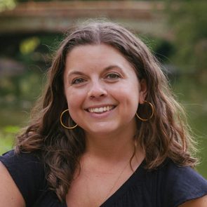 A woman smiles warmly, facing the camera. She has shoulder-length, wavy hair and wears hoop earrings, with a lush, green background suggesting a natural setting.