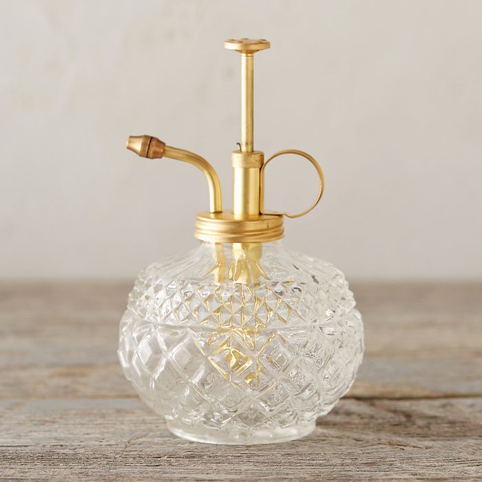 A glass spray bottle with a gold nozzle releases liquid, placed on a rustic wooden surface against a soft, neutral background.