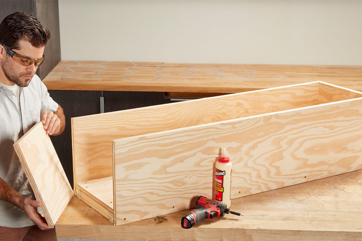 A man assembles a wooden box, holding a side panel, in a workshop with a wooden countertop and tools nearby.