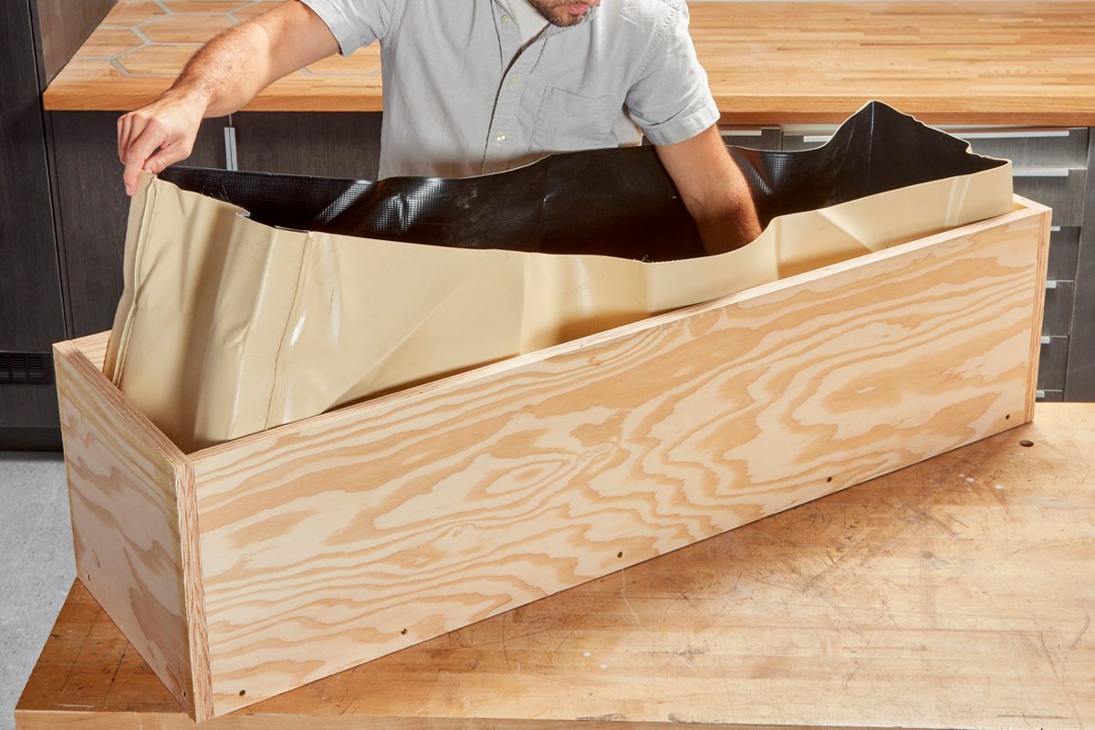 A person is placing a flexible liner into a wooden planter box on a workbench in a kitchen-like setting.
