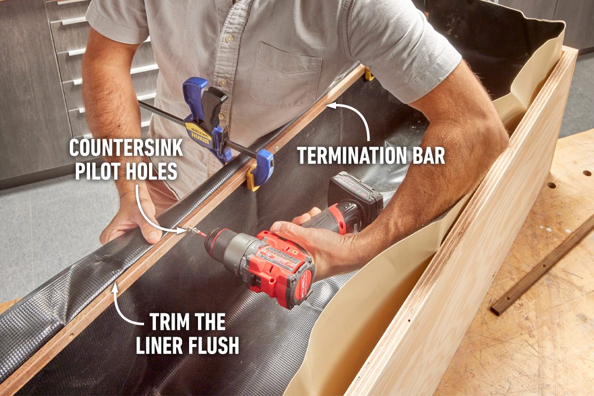 A person uses a drill to attach a termination bar while clamping a liner in place on a wooden structure, surrounded by tools and work surfaces.