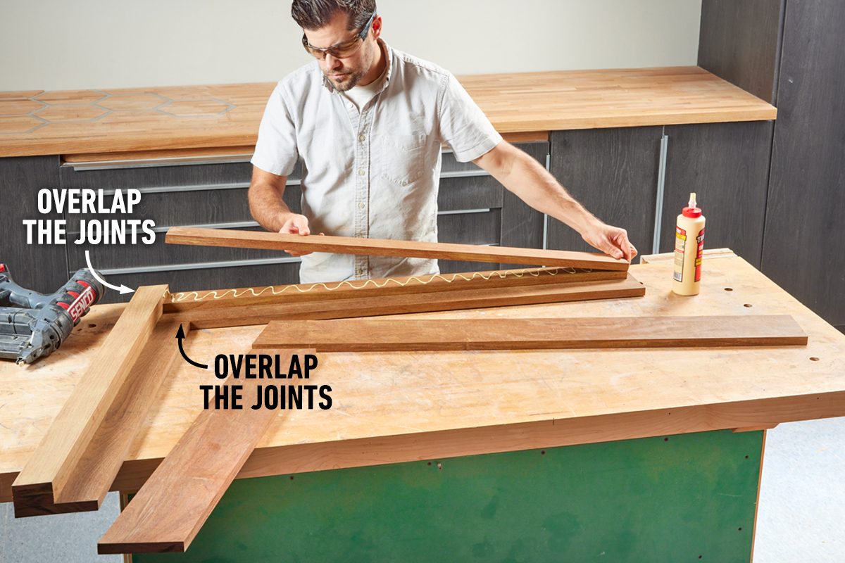 A person uses wood glue to assemble wooden pieces, overlapping the joints, on a workbench with safety glasses in a well-lit workshop.