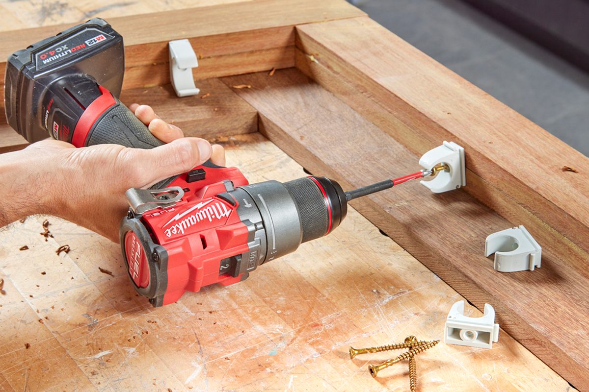 A drill is driving a screw into wood while held in a hand. Nearby, screws and white brackets rest on a wooden surface.
