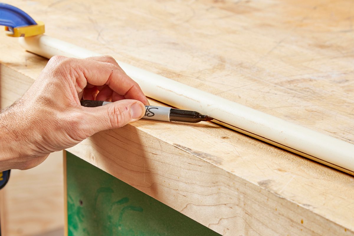 A hand holds a marker, drawing a line on a white pipe placed on a wooden work surface in a workshop environment.