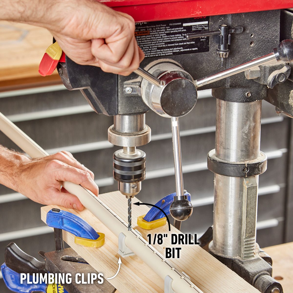 A person operates a drill press, using a 1/8" drill bit on a wooden piece secured with clamps, in a workshop setting.