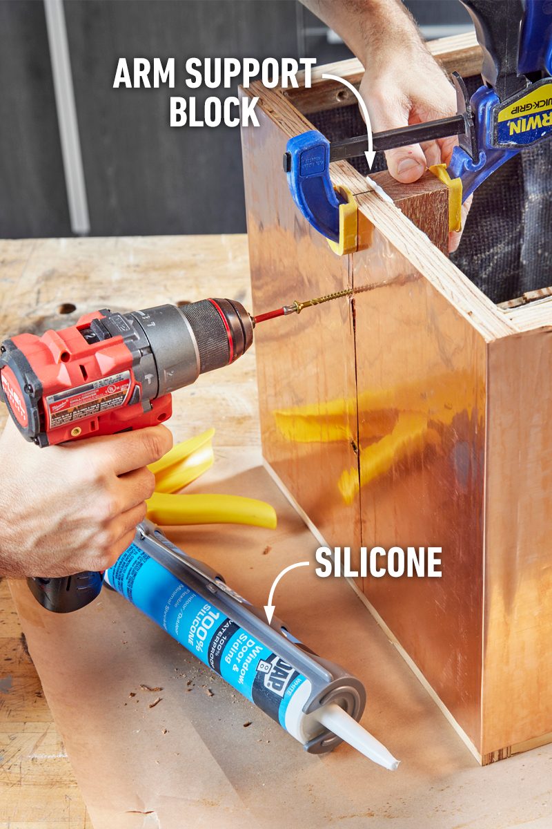 A drill is used to fasten wood while supported by a clamp, alongside silicone on a workspace covered with a protective sheet.
