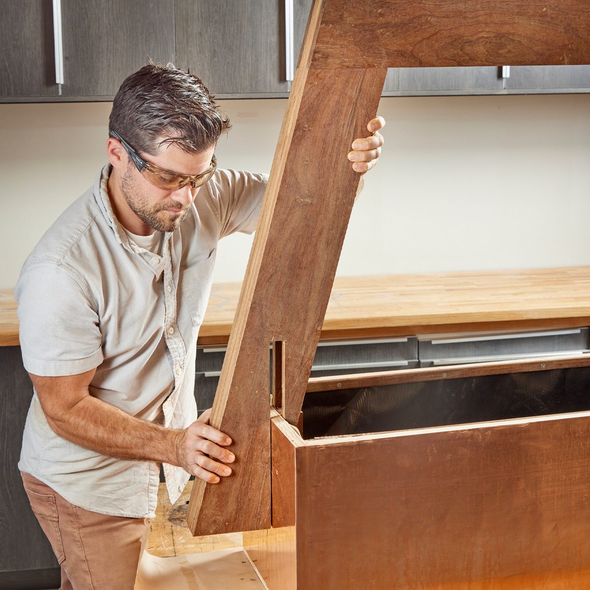 A man lifts a large wooden lid, adjusting it over a box in a modern workshop with wooden countertops and grey cabinetry.
