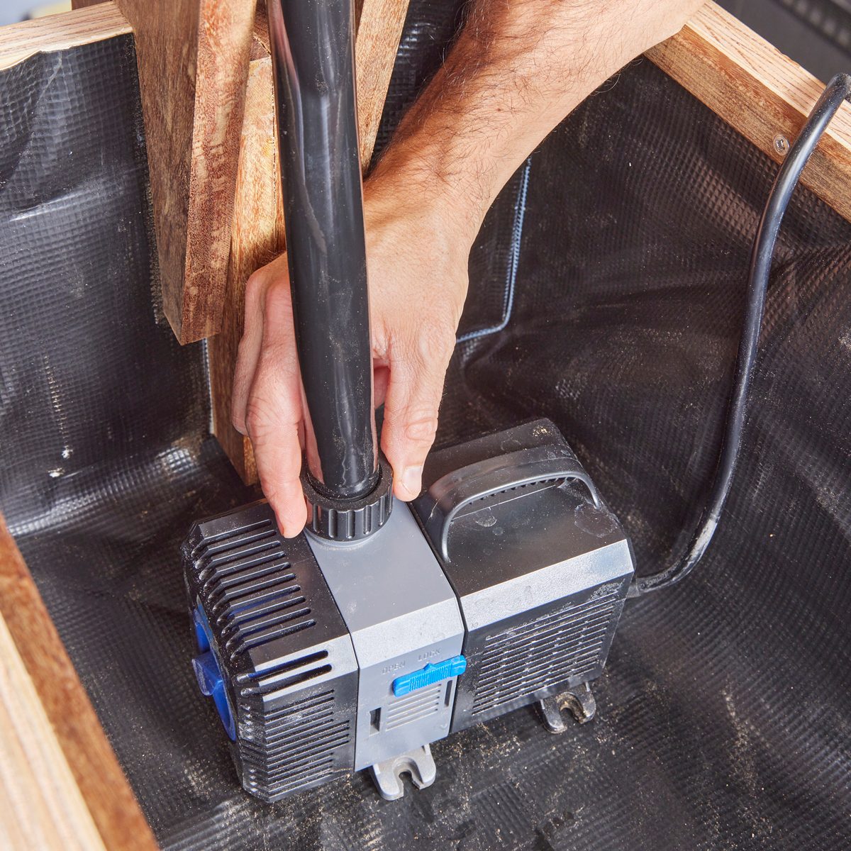 A person secures a black hose to a pump inside a wooden structure, which features a black, textured liner. The environment appears functional and utilitarian.