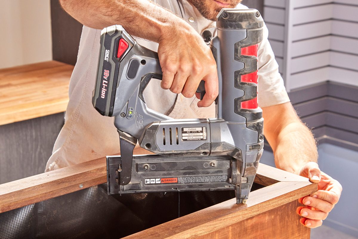 A person uses a nail gun to fasten wood pieces together on a work surface in a bright, modern environment.