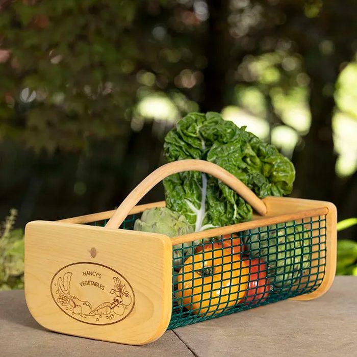 A wooden basket holds various vegetables, including leafy greens and fruits, resting on a surface with a blurred, natural backdrop.