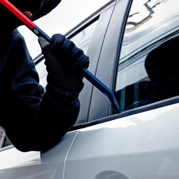 A person in a black hoodie uses a crowbar to pry open a car window in a street setting.