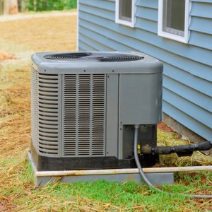 An air conditioning unit stands on a grassy area beside a light blue house, connected to pipes and electrical wiring, in a serene outdoor setting.
